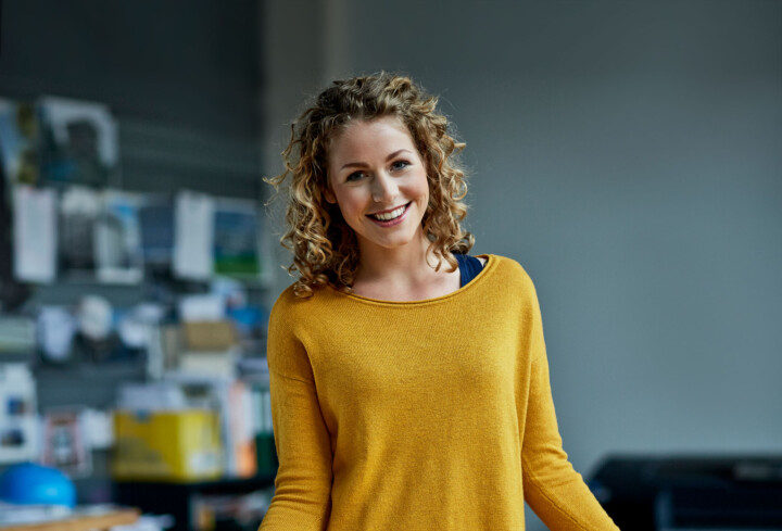 Jonge, blonde vrouw met gele trui kijkt lachtend inde camera. Op de achtergrond een kantoor met bouwtekeningen.